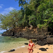 Woman in a red bikini standing on a sandy beach with rocky outcrops and greenery in the background.