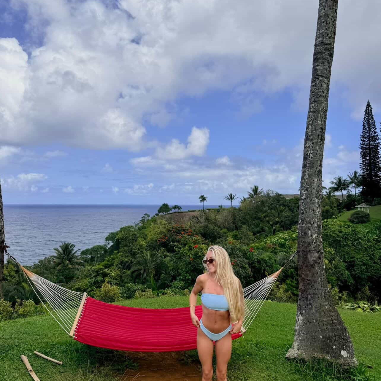 Woman in a blue bikini standing next to a red hammock with a tropical landscape in the background.