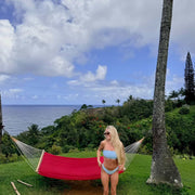 Woman in a blue bikini standing next to a red hammock with a tropical landscape in the background.