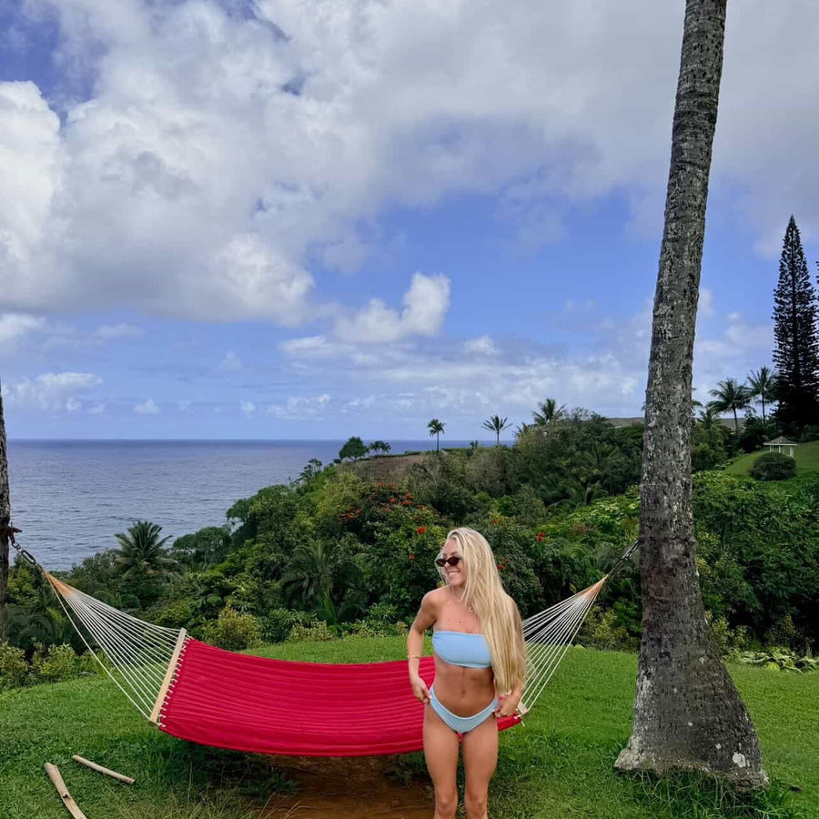 Woman in a blue bikini standing next to a red hammock with a tropical landscape in the background.